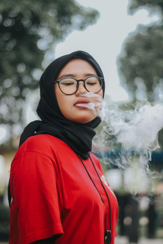 A woman in glasses and a red shirt smokes, showcasing a Packwoods x Runtz disposable vape in her hand.